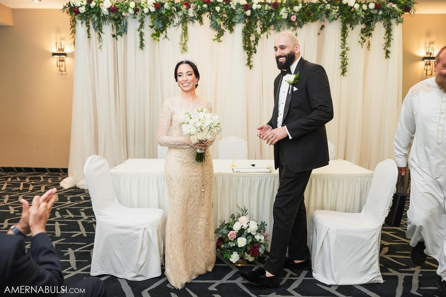 Wedding couple standing under floral arch with white chairs and elegant decor.