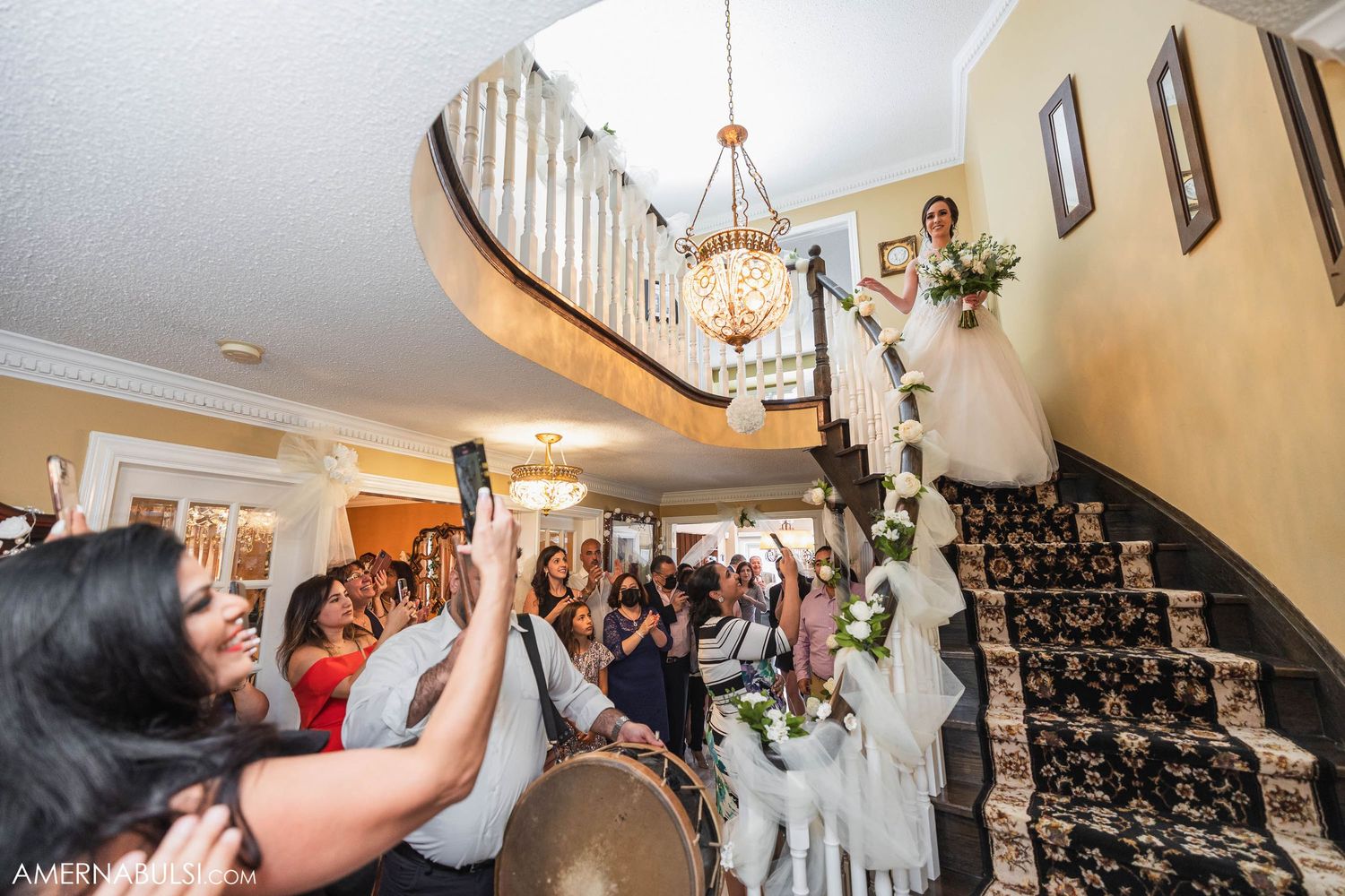 Wedding guests cheer as the newlyweds make their grand entrance down an elegant curved staircase.