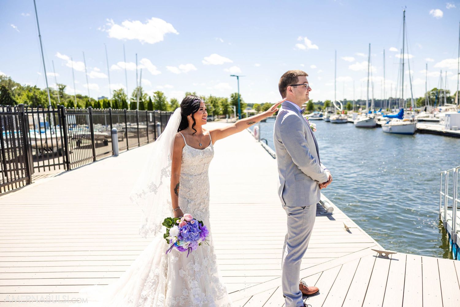 A couple shares their first look moment on a marina dock surrounded by sailboats on a sunny day.