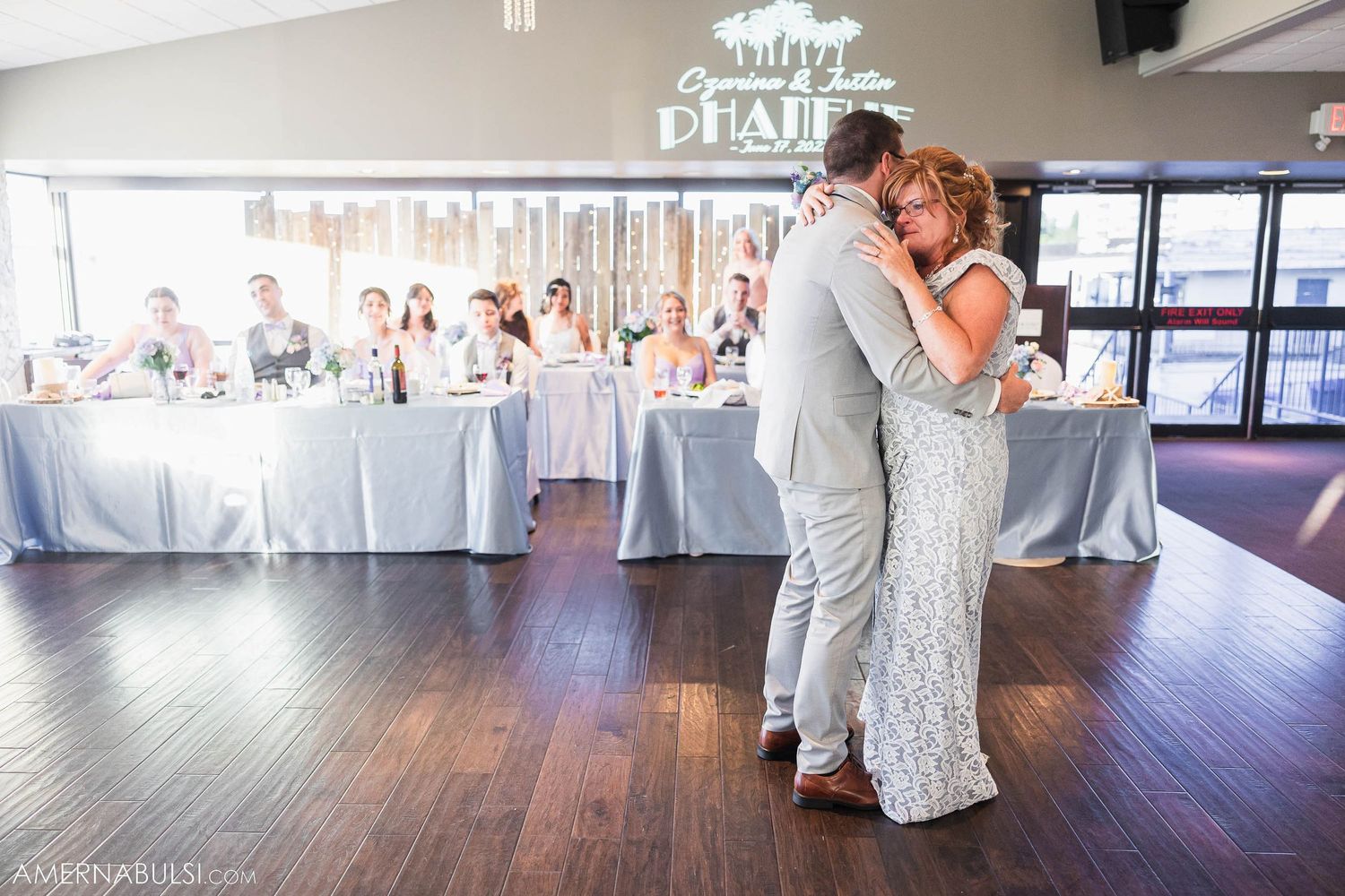 Newlyweds share first dance on wooden floor with decorated reception tables in background.