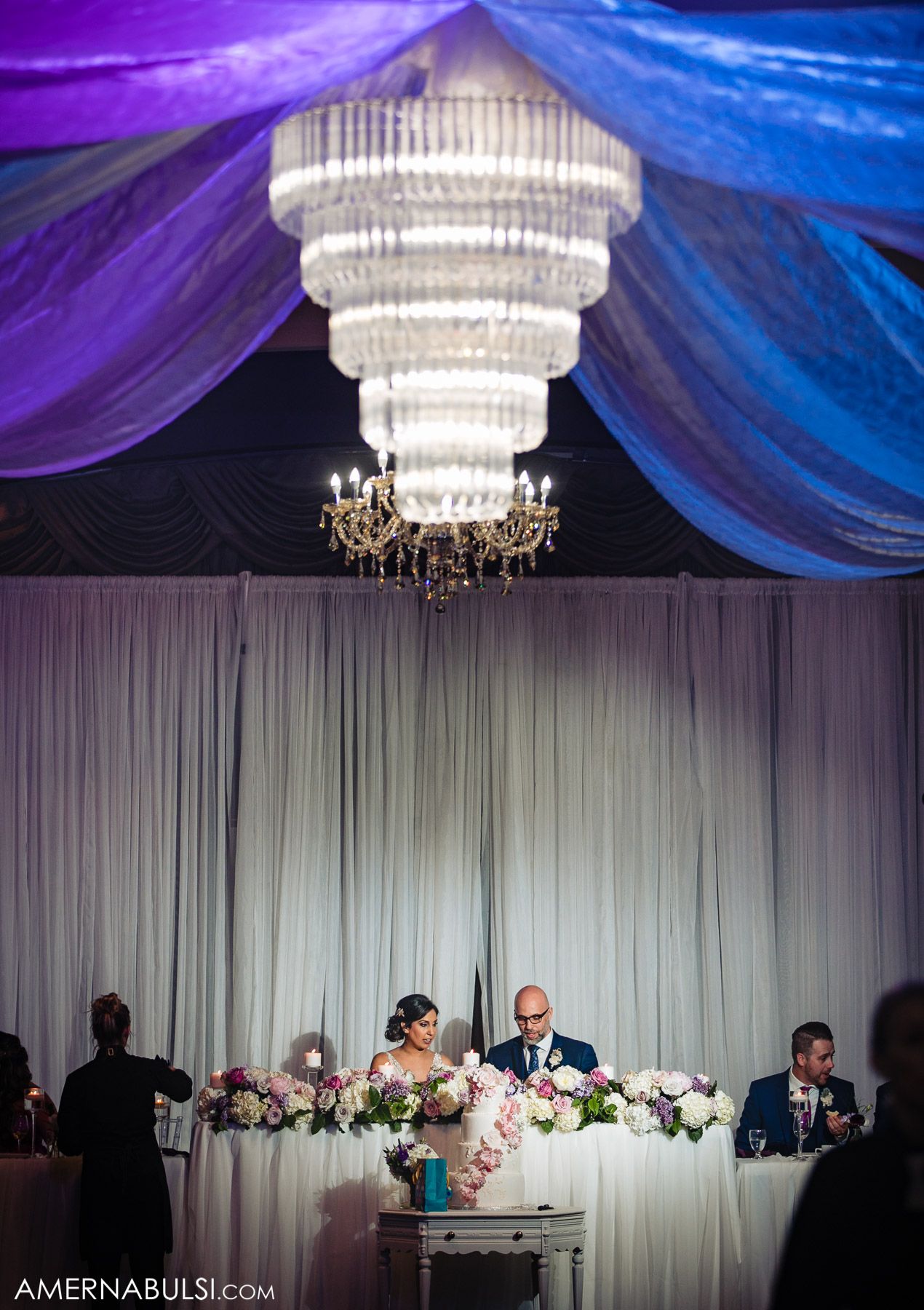 An elegant chandelier hangs above a sweetheart table draped with flowers and blue fabric decor.