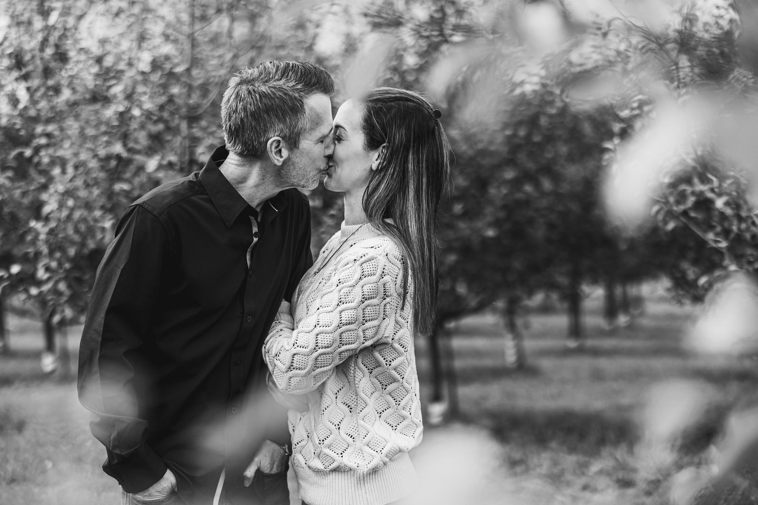 A romantic couple embraces in a dreamy black and white outdoor setting with blooming trees in the background.