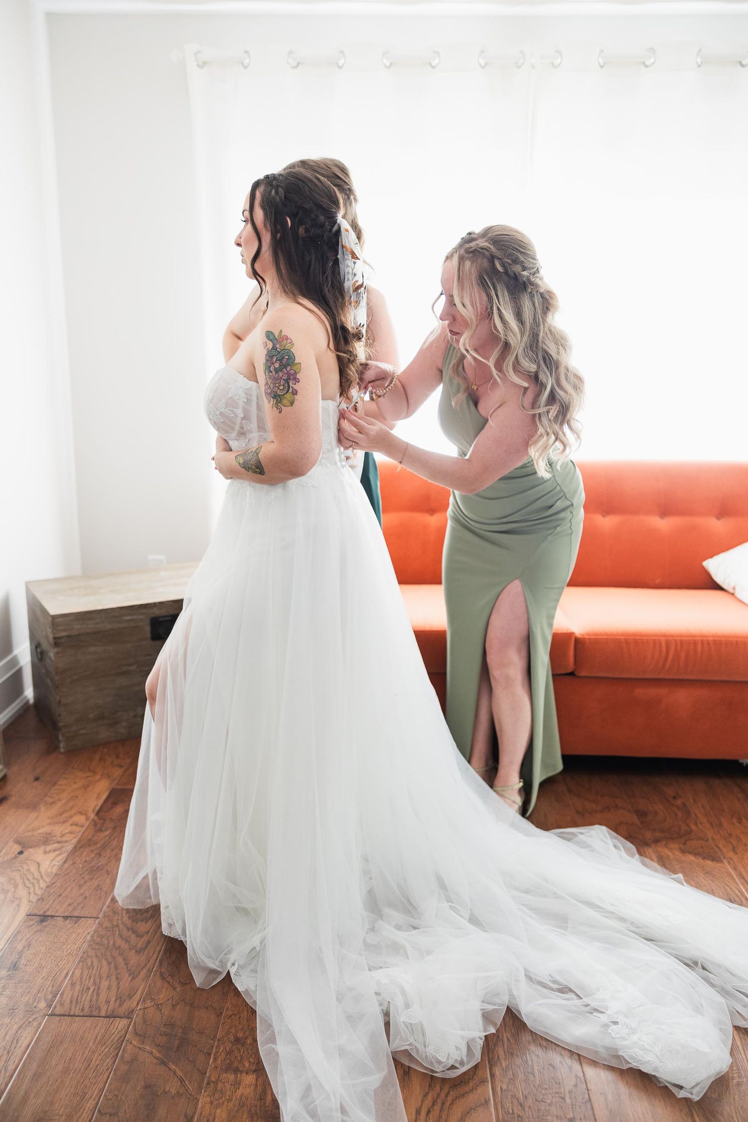 A bridesmaid in sage green helps adjust the bride's gown in a room with orange couch.