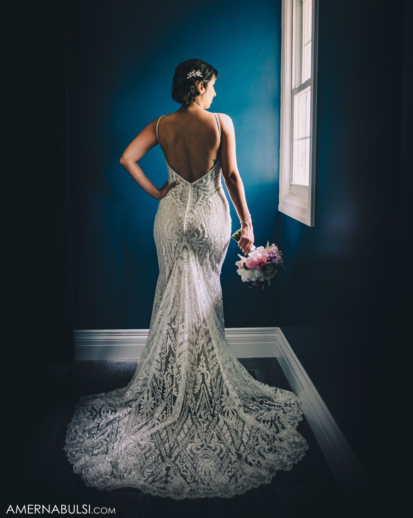 A stunning lace wedding dress with a dramatic train photographed against a deep blue wall in moody lighting.
