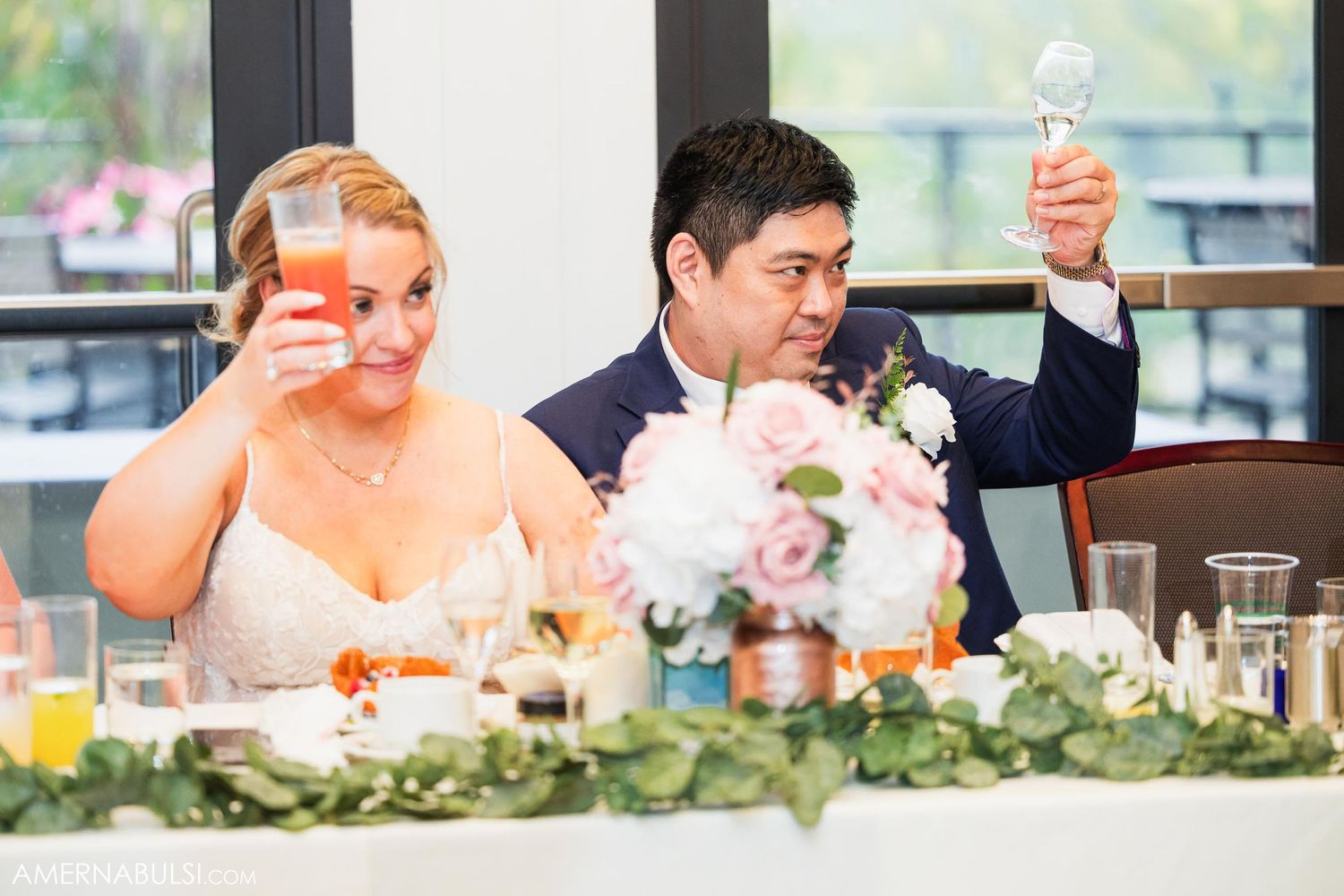 Newlyweds at their reception table raise glasses in a celebratory toast among floral decorations.