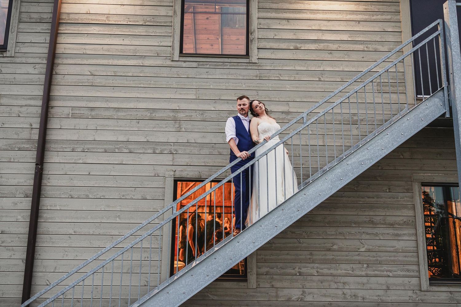 A couple poses on an outdoor staircase against weathered wooden siding at their wedding venue.