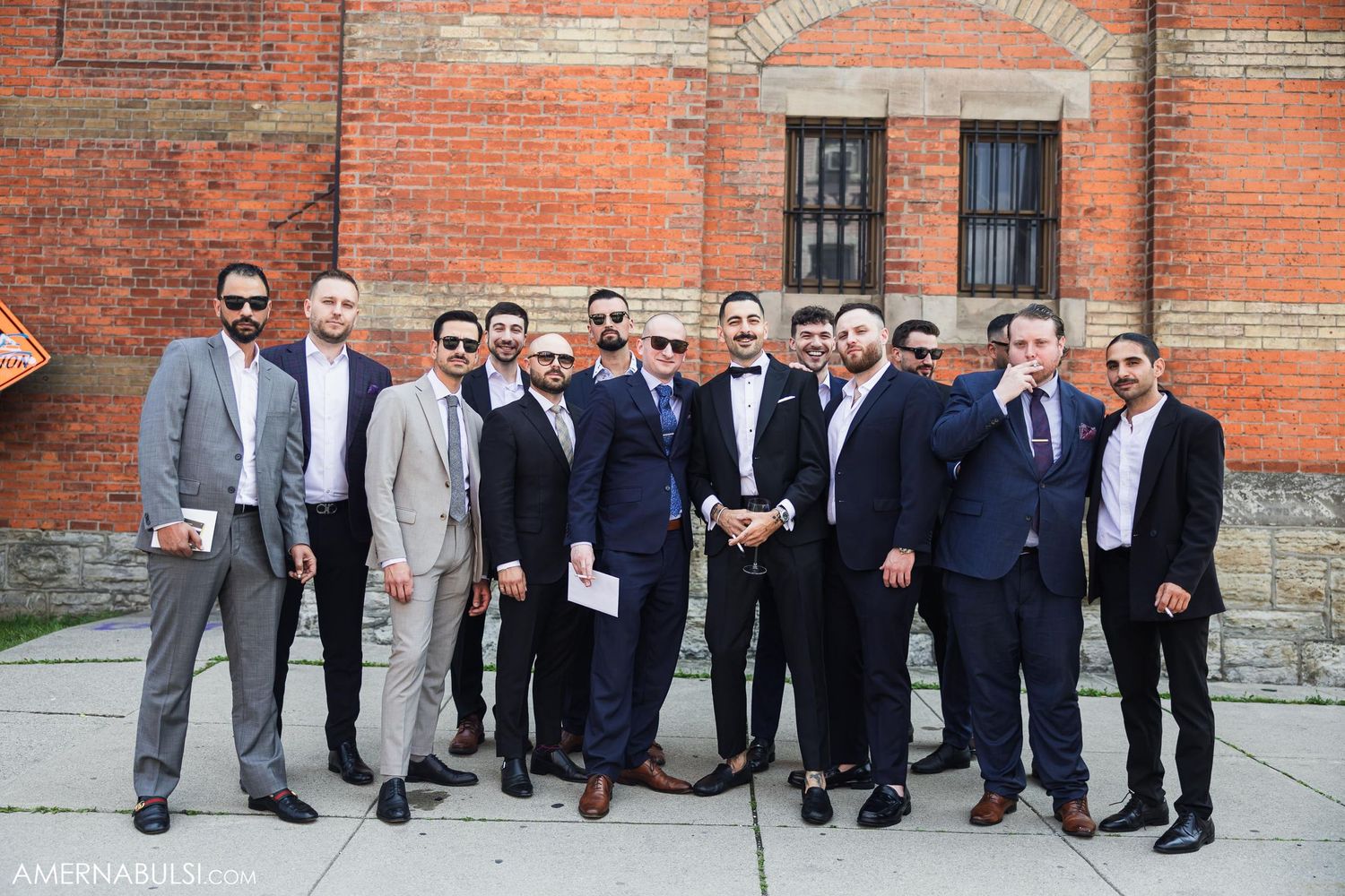 Group of men in formal attire pose together against rustic brick building exterior.