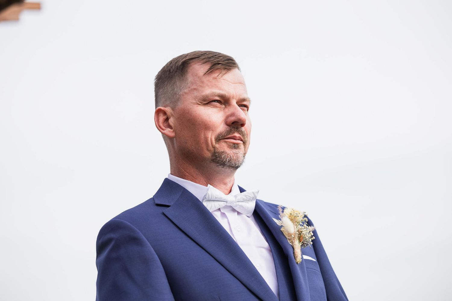 Close-up view of groom in navy suit with boutonniere against white sky.