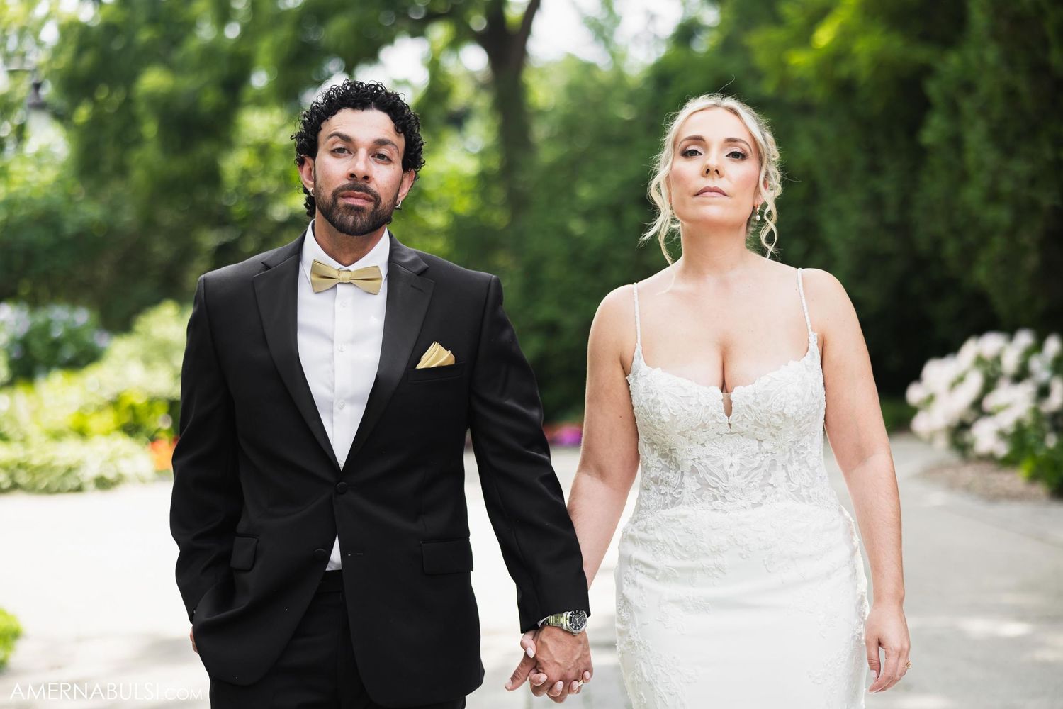 Newlyweds walk hand in hand along garden path in formal wedding attire.