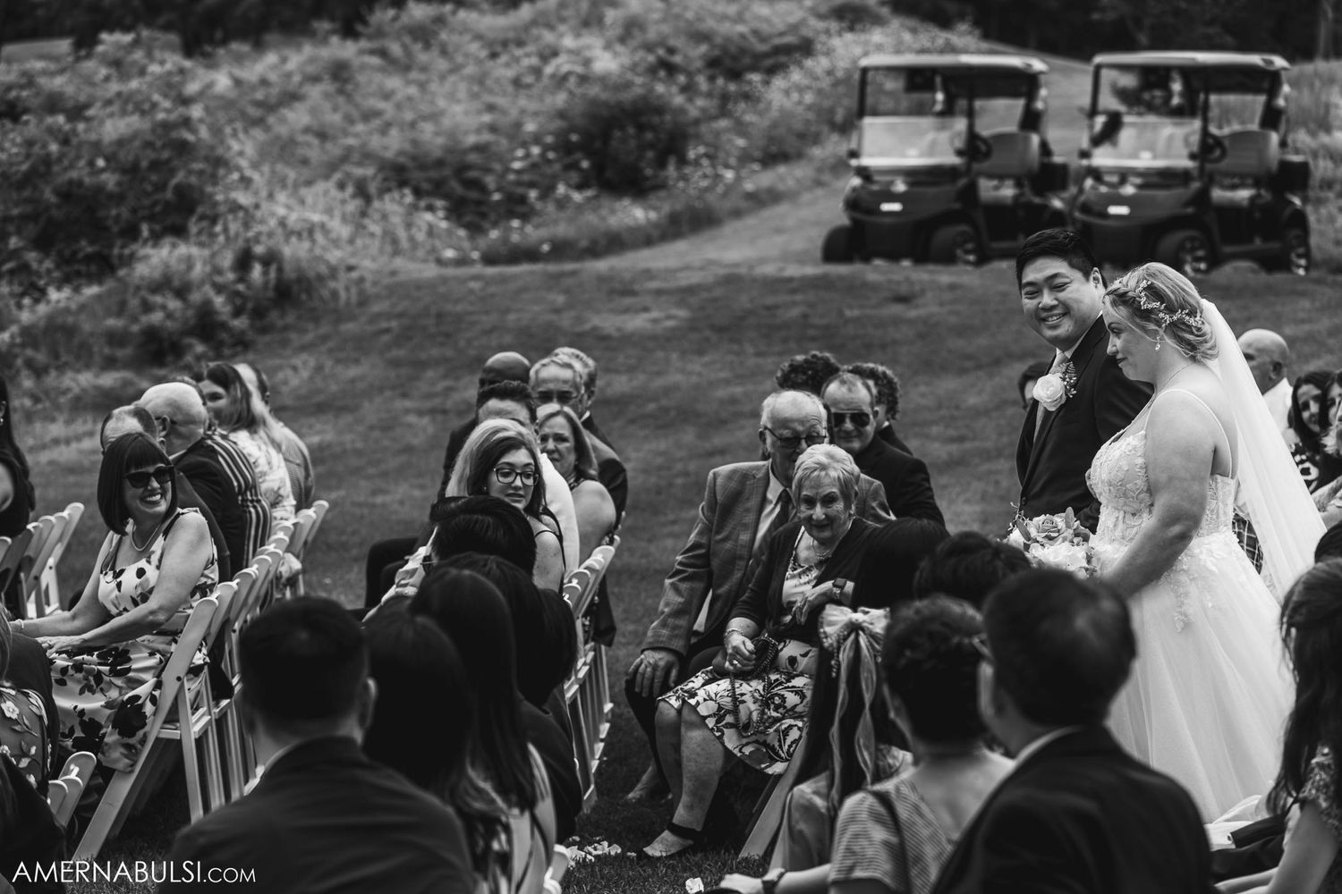 Black and white photo of wedding guests watching ceremony with golf carts parked on hillside.