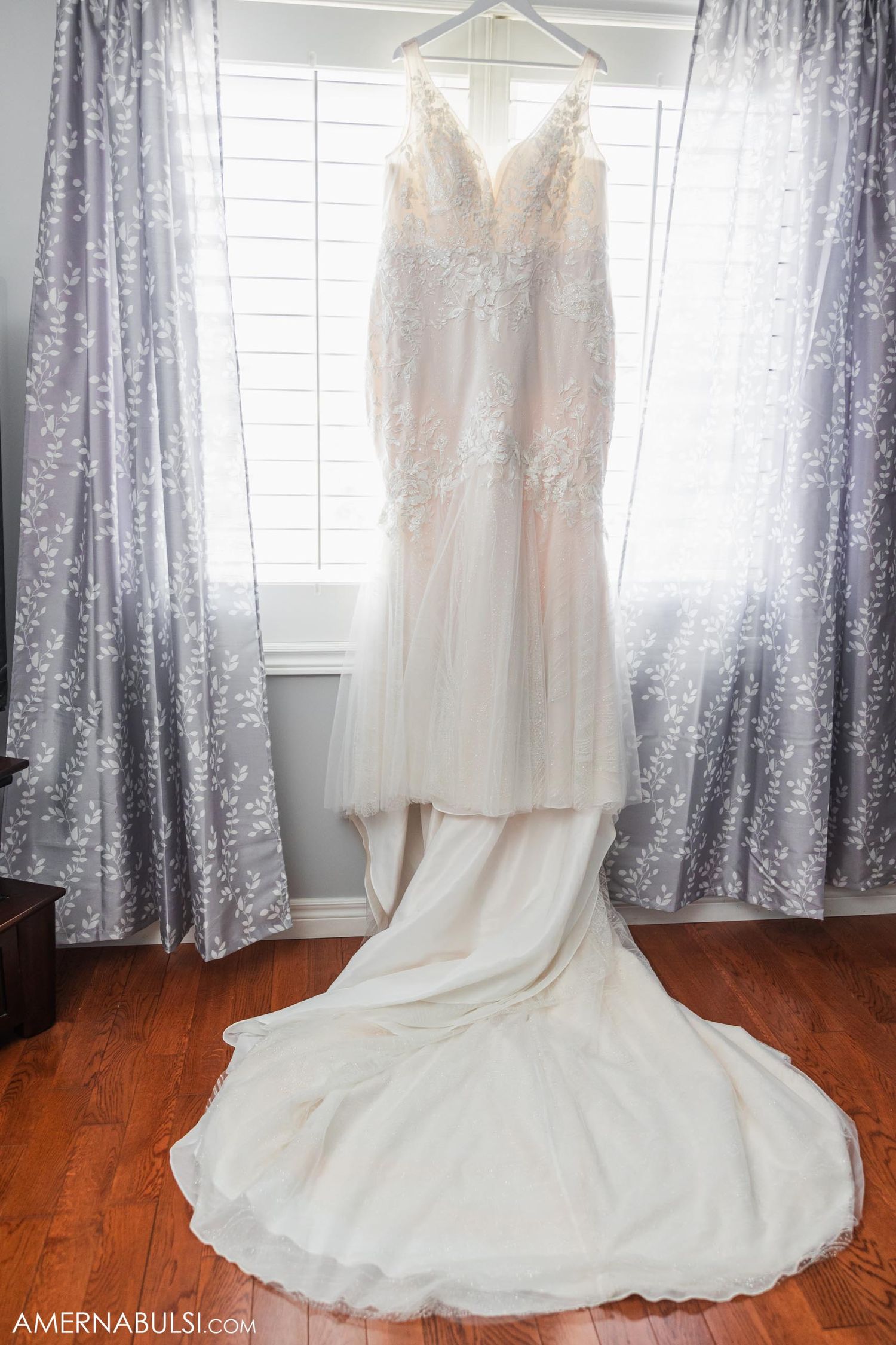 Elegant white wedding dress with lace details hanging in front of silver curtains.
