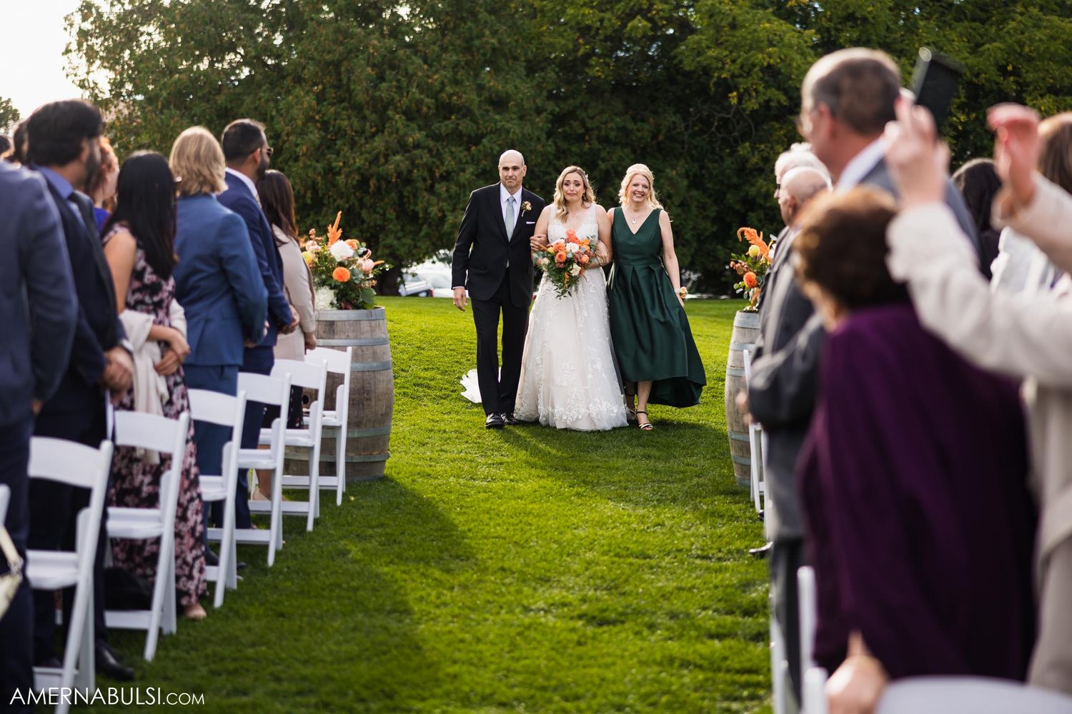 Wedding guests line white chairs along a grassy aisle as a wedding party processes.
