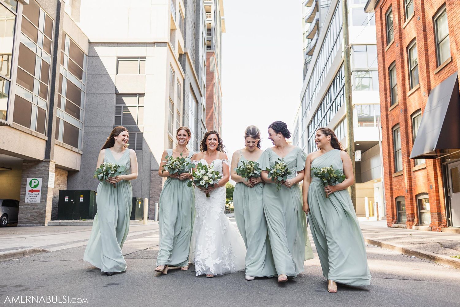 Wedding party walking together in sage green outfits holding bouquets in urban setting.