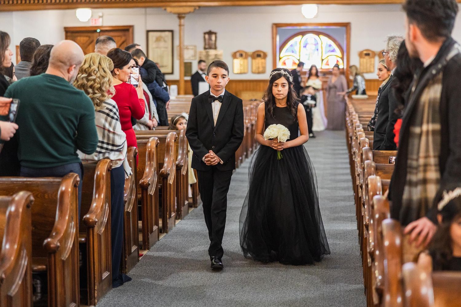 Wedding party members in formal black attire walk down church aisle during ceremony.