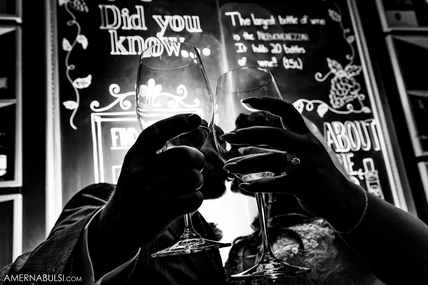 Black and white silhouette of hands toasting glasses against illuminated chalkboard.