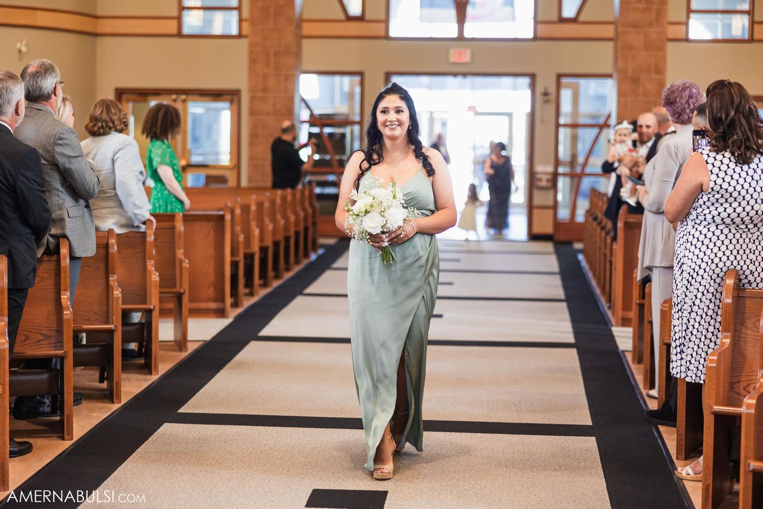 A bridesmaid walks down the church aisle carrying a white bouquet during a wedding ceremony.