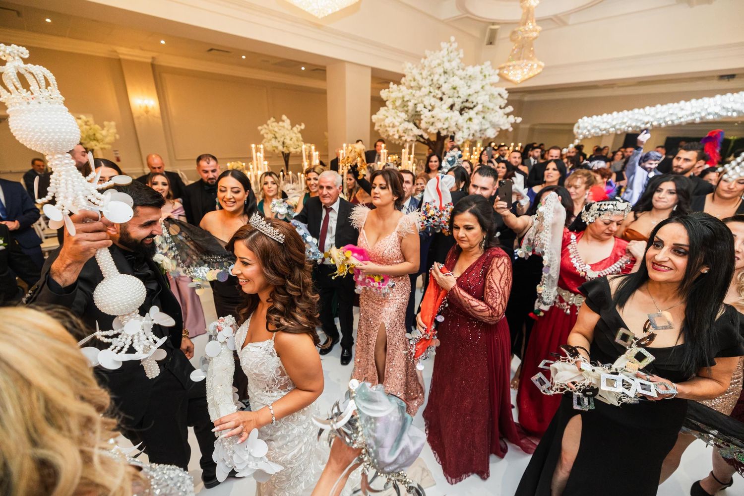 Wedding guests celebrate in an elegant ballroom with white floral arrangements and chandeliers.