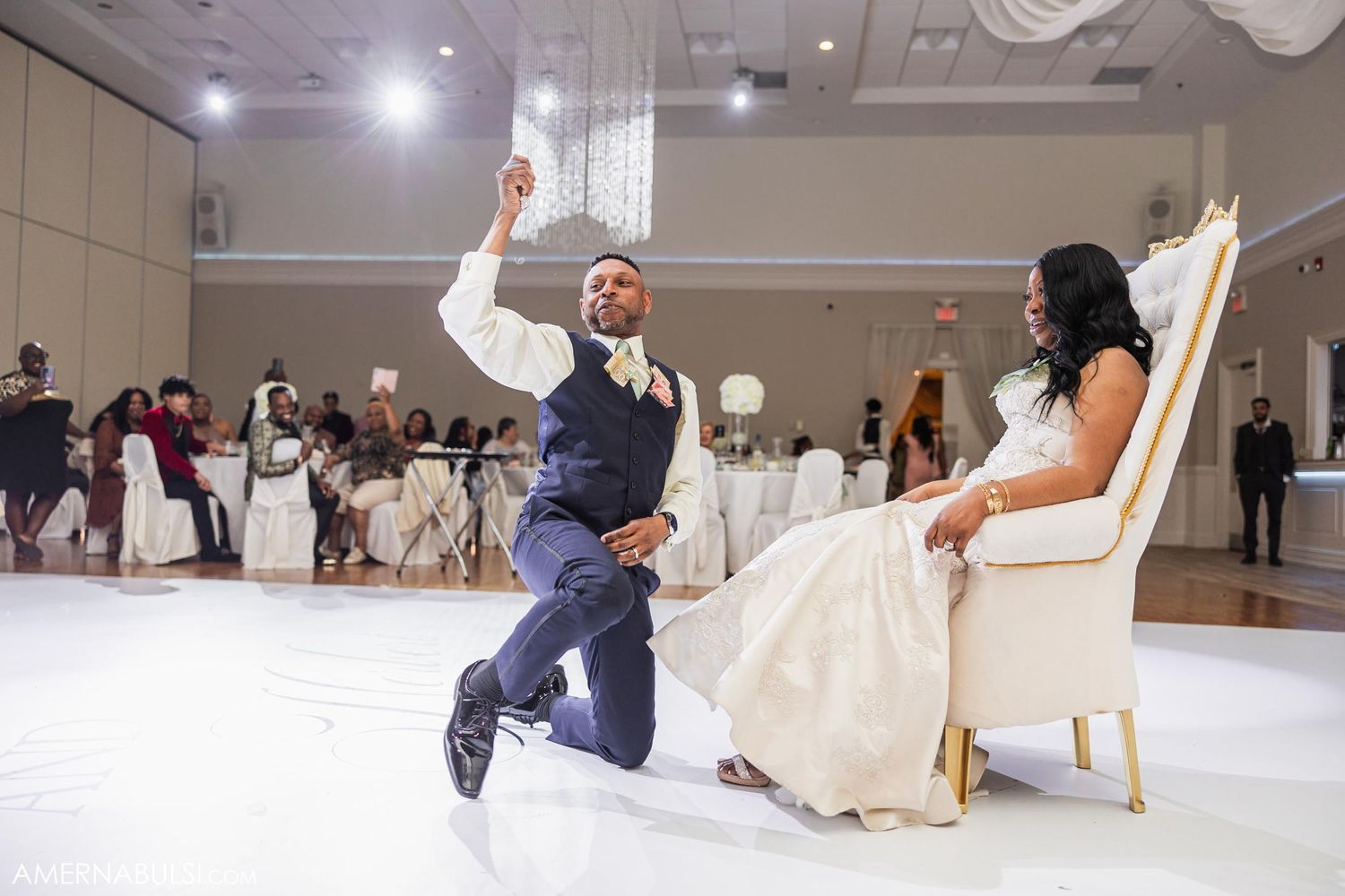 A groom performs an energetic dance move during a wedding reception while the bride watches.