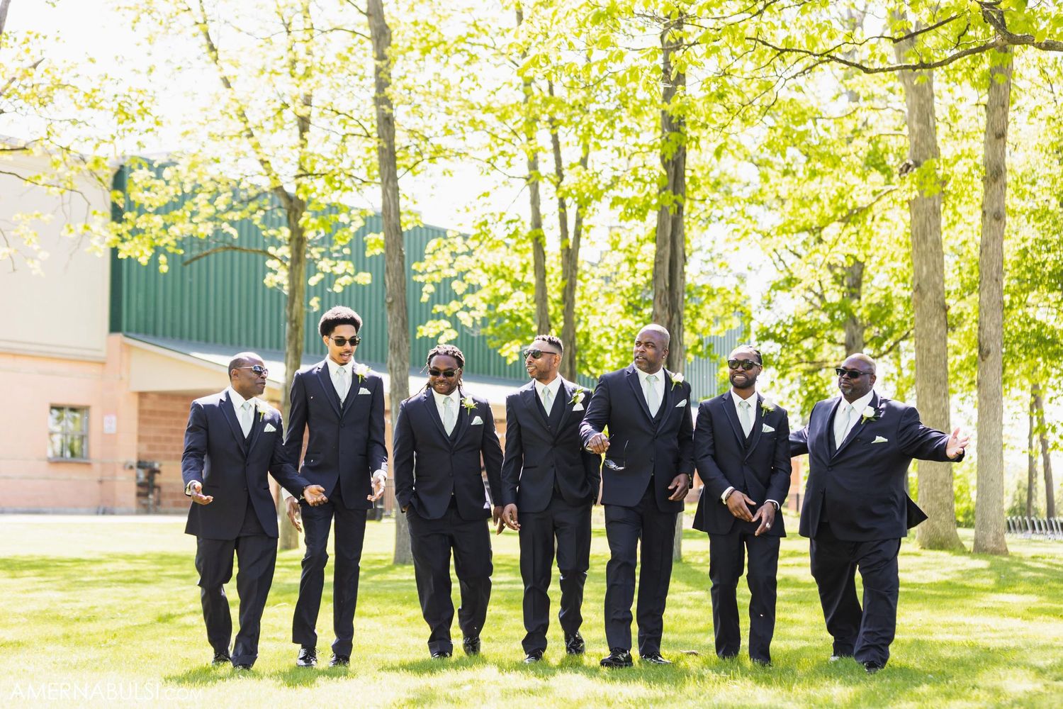 Wedding party groomsmen in black suits walking together on a sunny day through a garden setting.