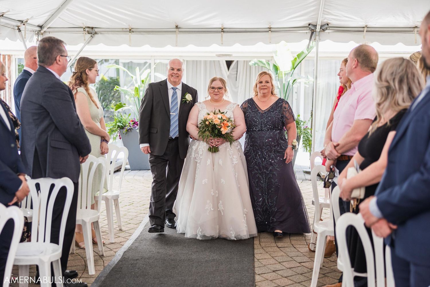 Wedding ceremony processional in a tent with white chairs and guests lined along the aisle.
