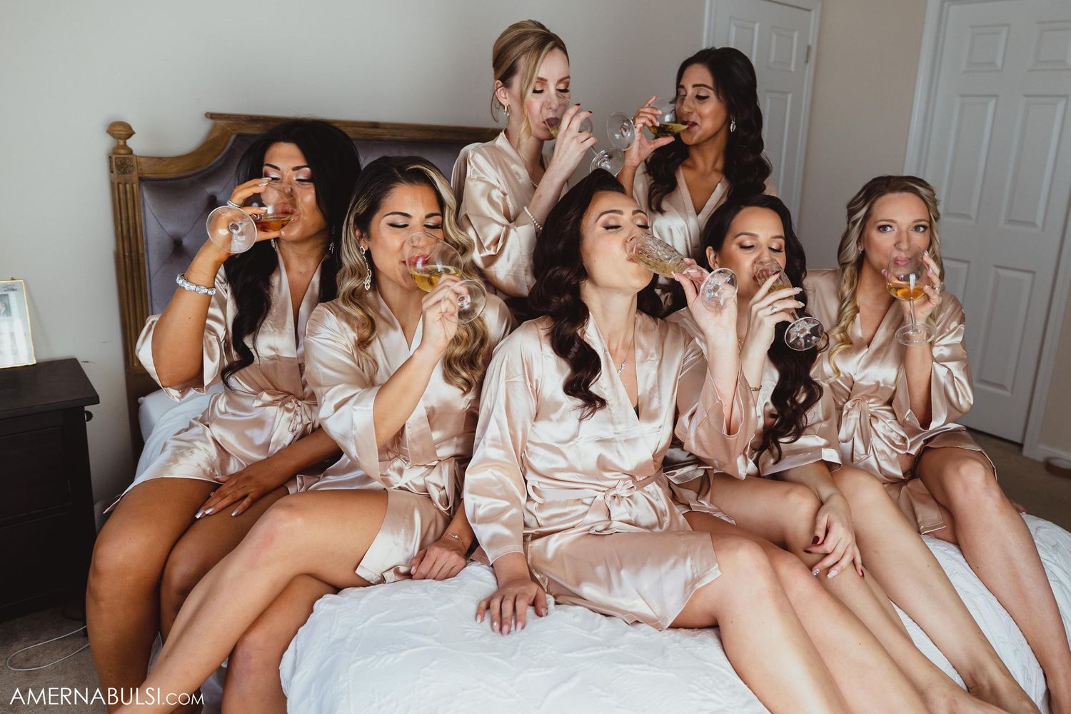 Bridesmaids in silk robes pose together on a bed during wedding morning preparations.
