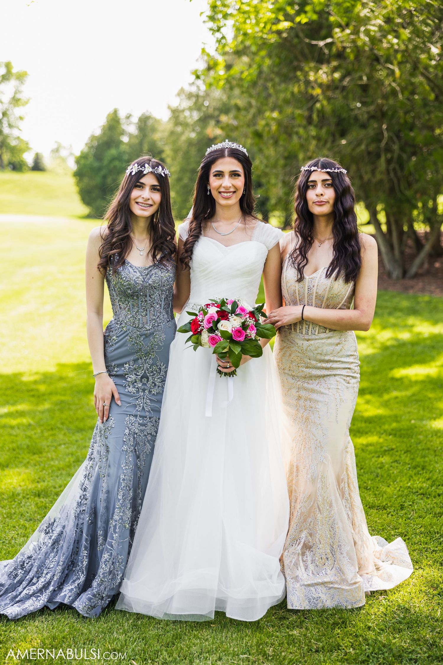 Three bridesmaids pose in elegant gowns for formal wedding photo on lush green lawn.