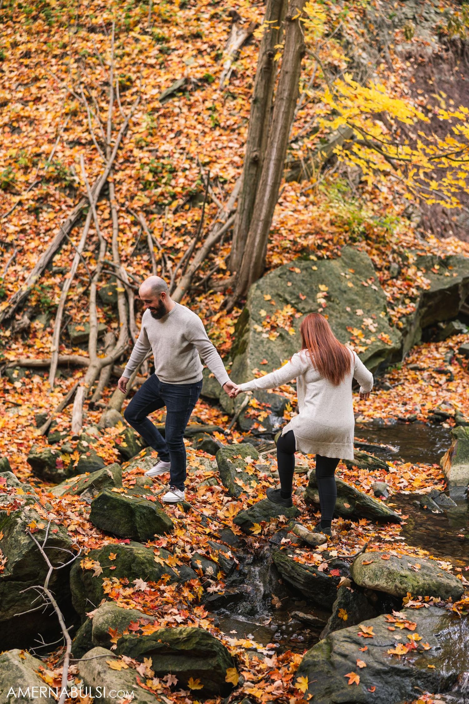Couple carefully crosses stream on rocks amid fall foliage.