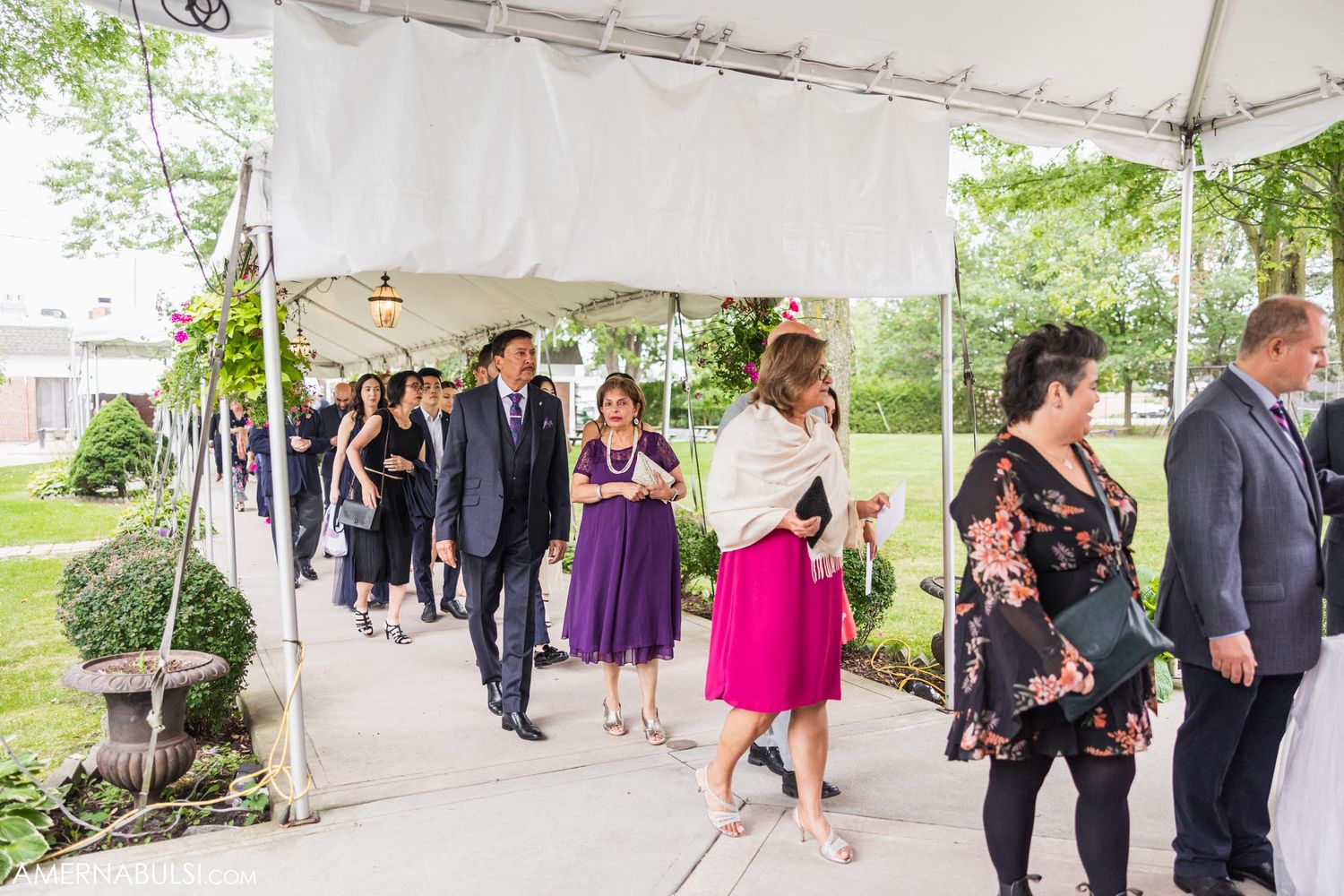 Wedding guests walk under a white tent decorated with greenery during a reception entrance.