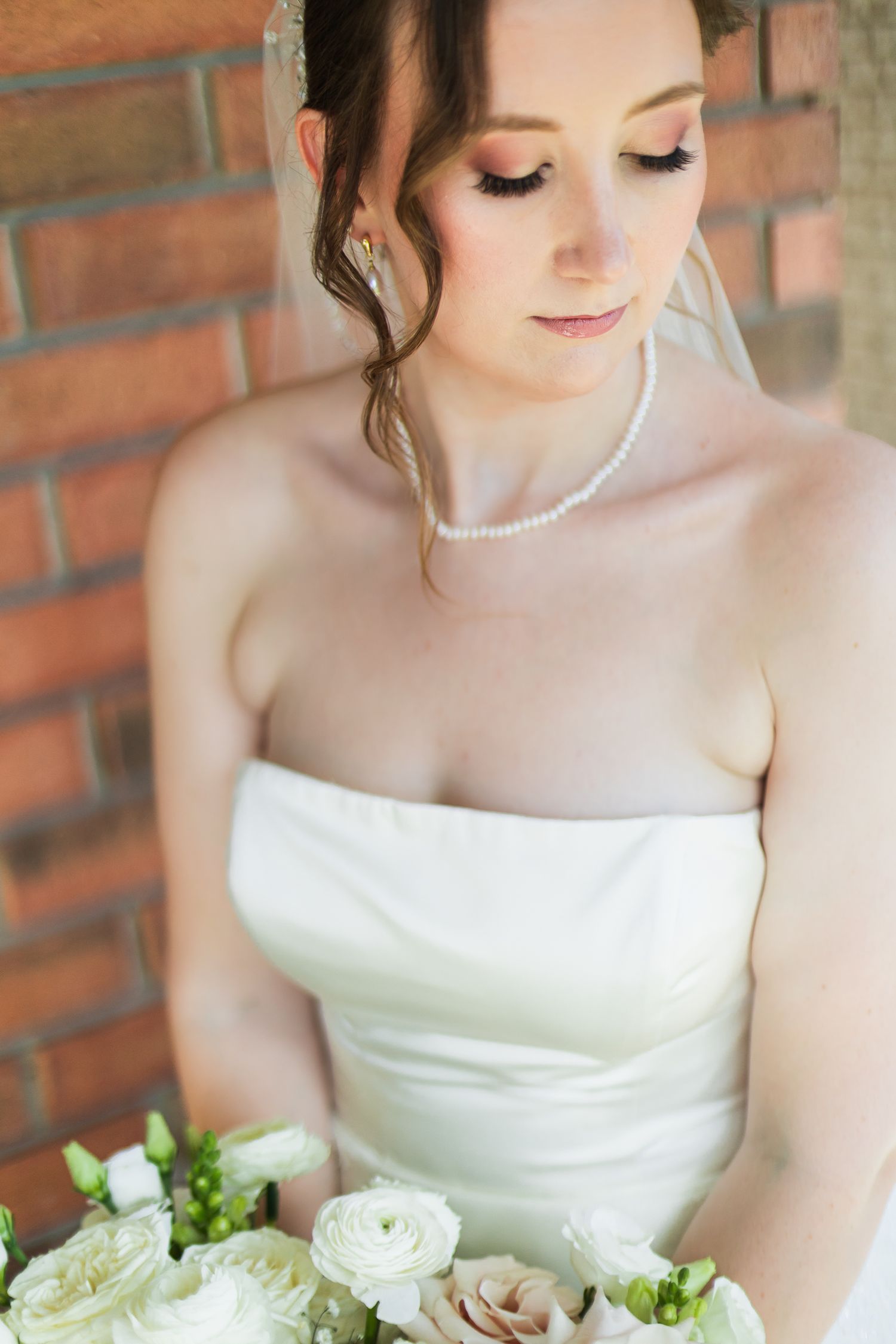 Elegant bride in strapless white gown wearing pearl necklace holds white bouquet against brick wall.