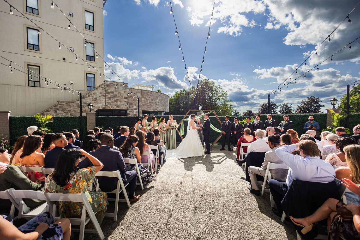 Outdoor wedding ceremony on a rooftop patio with guests seated in white chairs under string lights and a blue sky.