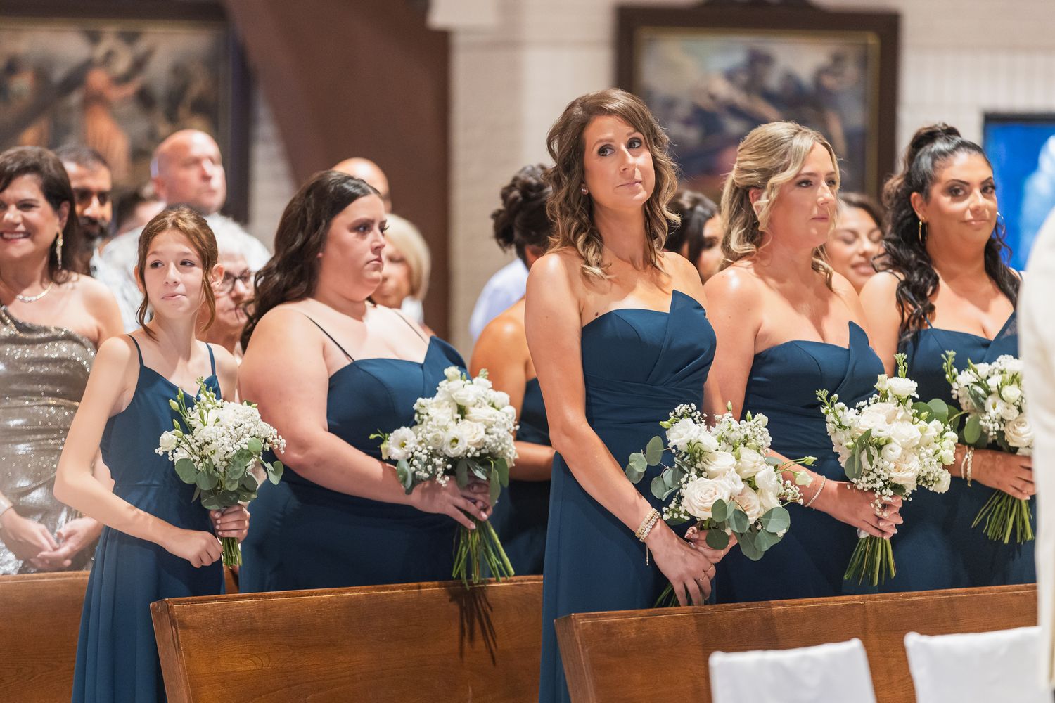 Bridesmaids in navy blue dresses holding white bouquets stand in a church aisle during a wedding ceremony.