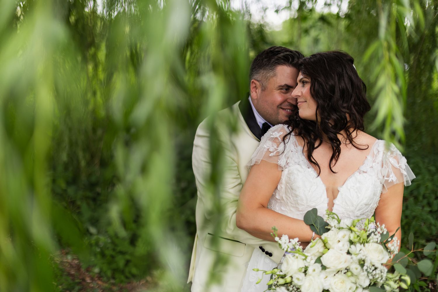 Bride and groom share a romantic moment nose-to-nose surrounded by lush green weeping willow branches.