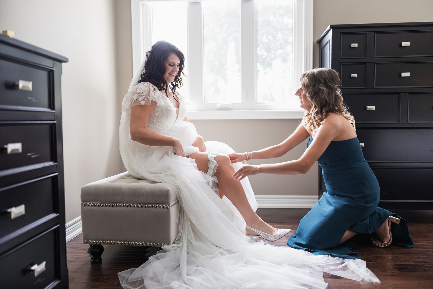 Bridesmaid in teal dress helps bride with her garter while she sits in a white lace wedding gown.