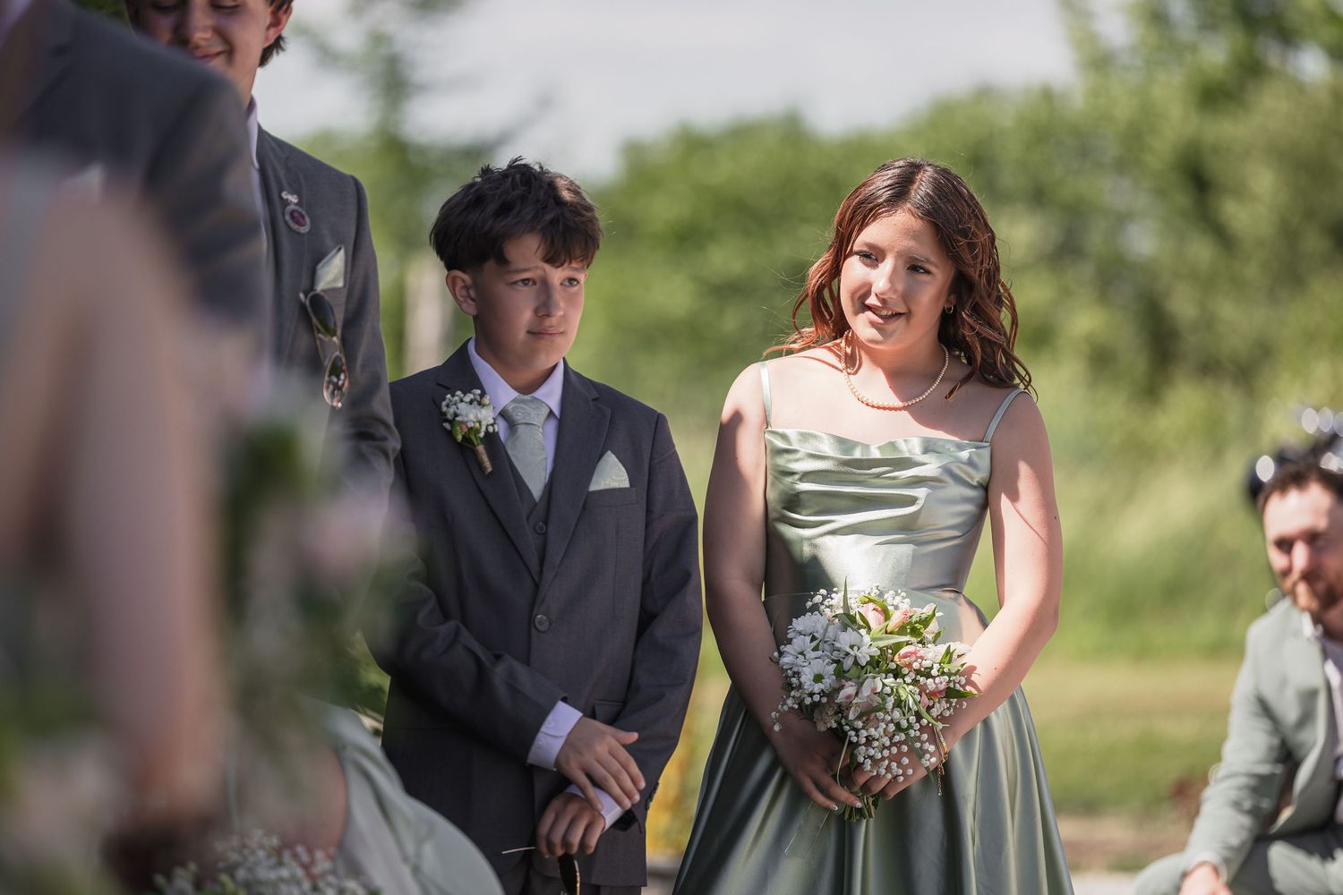 Bridesmaid in sage green dress smiles warmly during outdoor wedding ceremony.