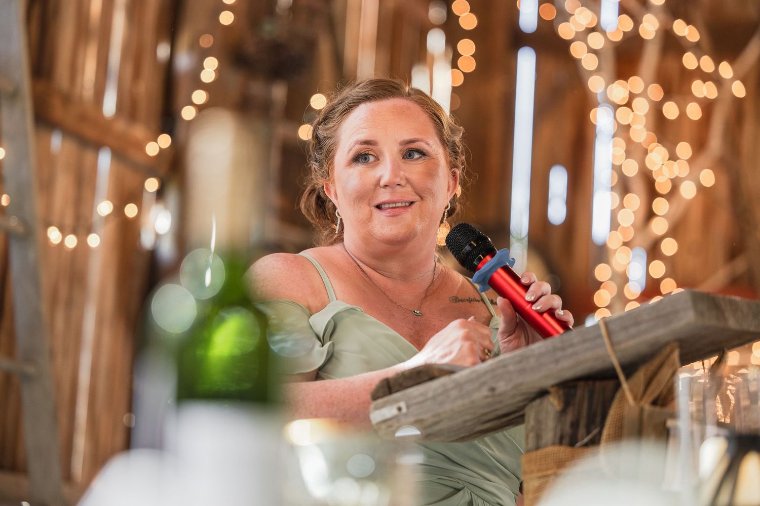 A bridesmaid in a sage green dress holds a microphone while giving a heartfelt speech at a rustic barn wedding reception.