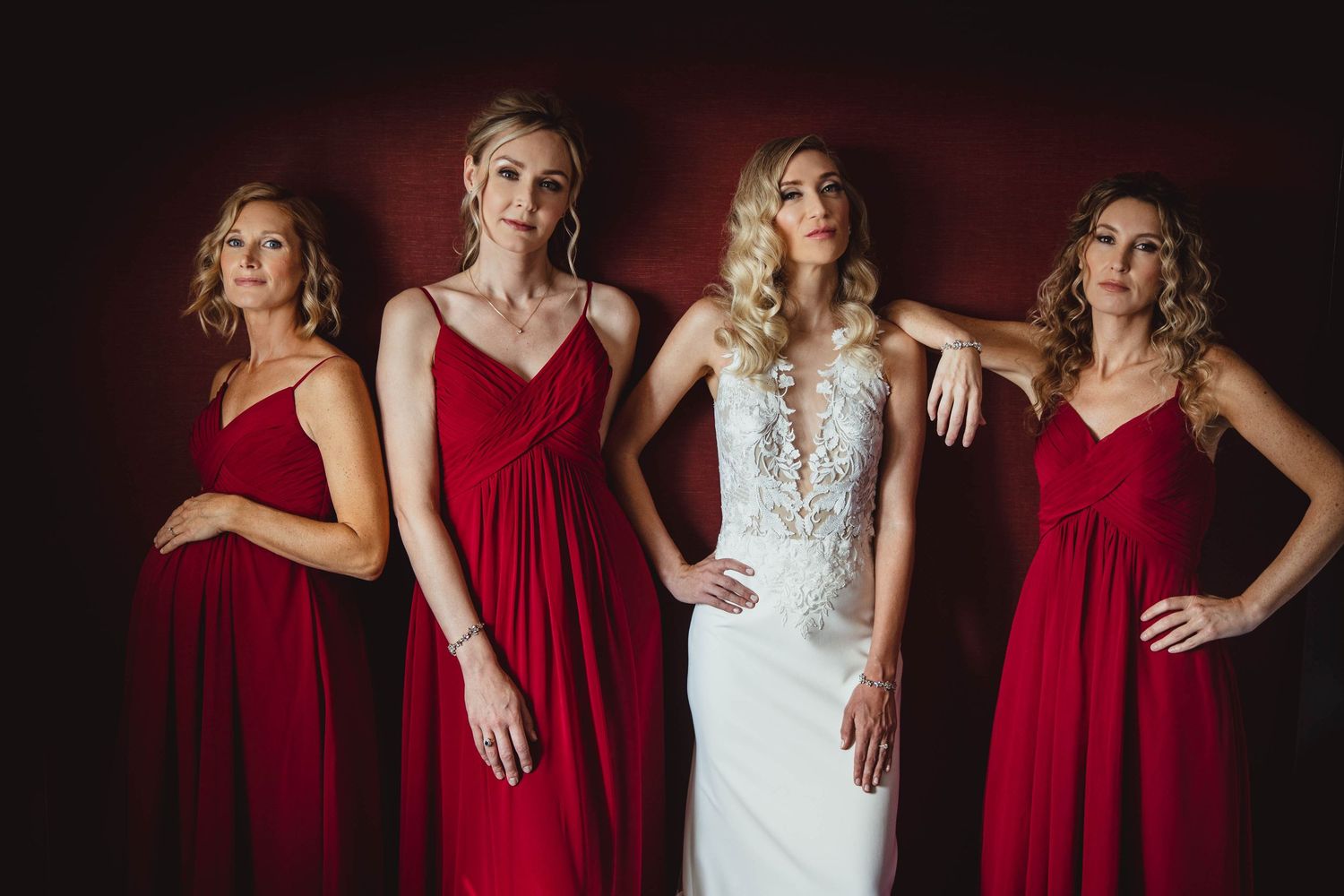 Bridal party poses against deep red backdrop wearing crimson dresses.