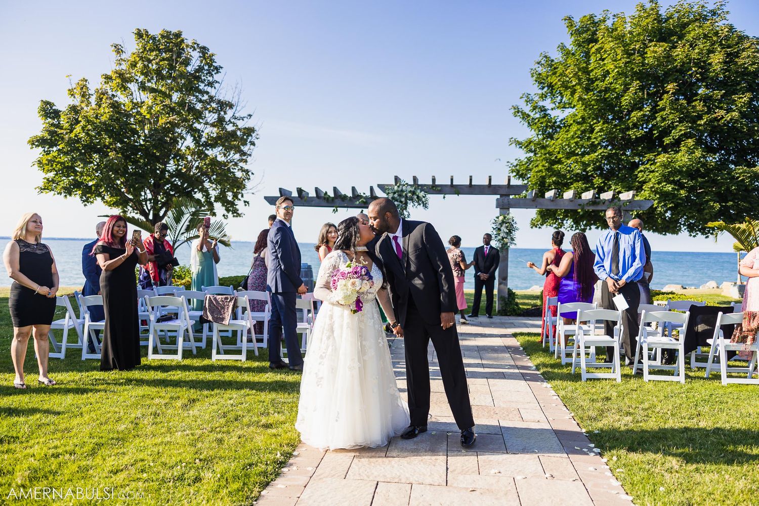 A wedding ceremony takes place on a scenic outdoor patio with a pergola overlooking water on a sunny day.