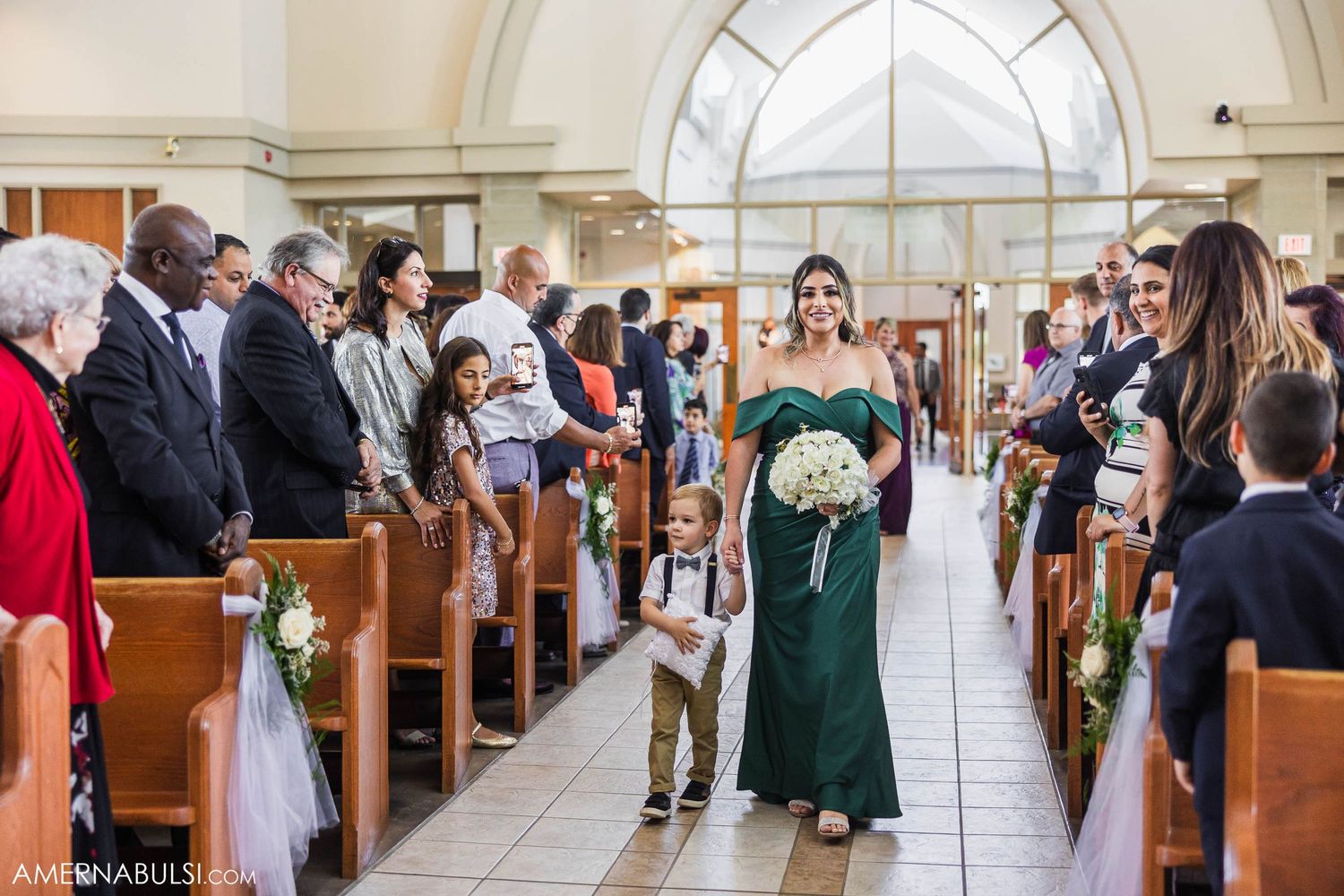 A bridesmaid in emerald green walks down the church aisle holding a white bouquet during a wedding ceremony.