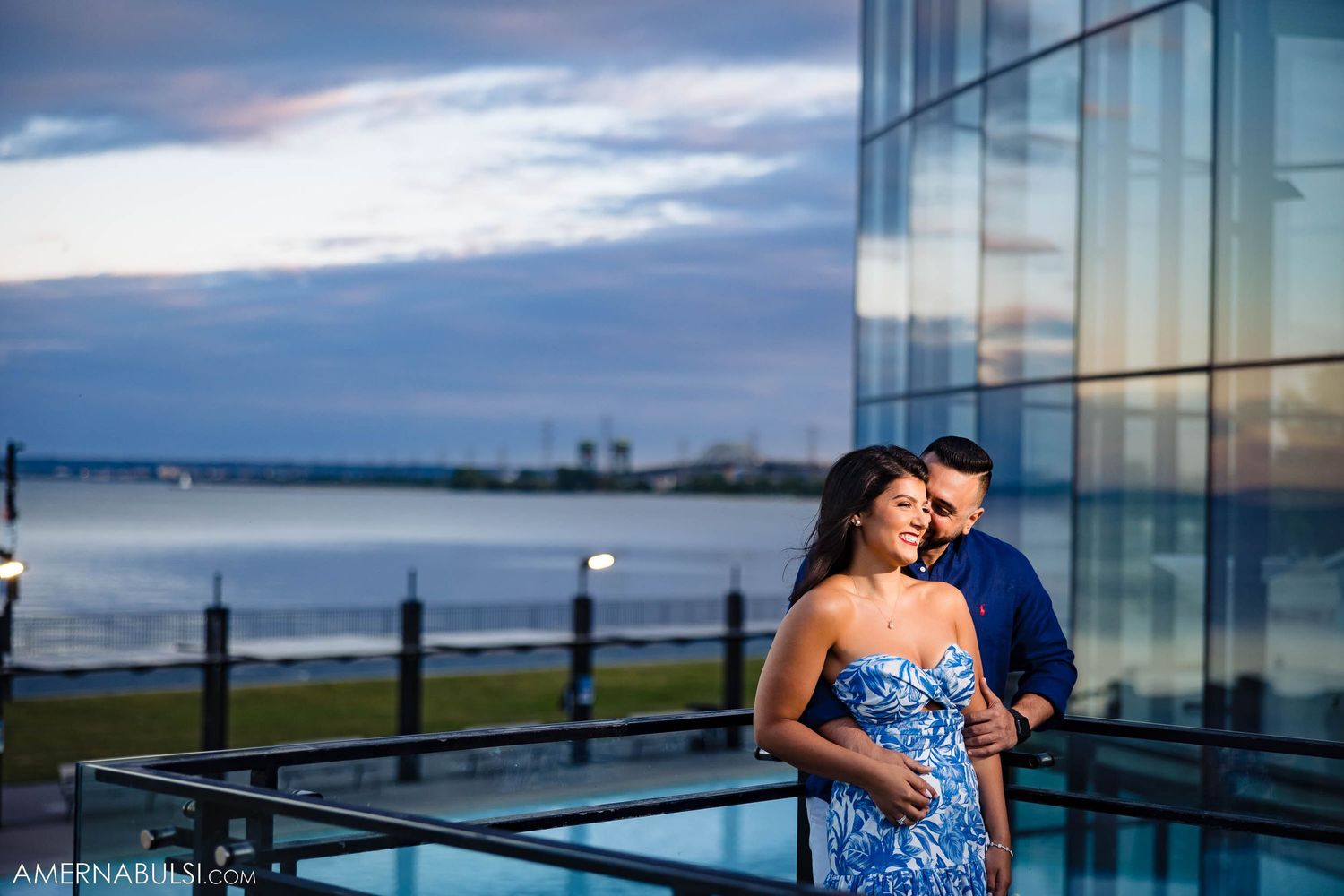 Couple embraces near glass building with waterfront view during blue hour.