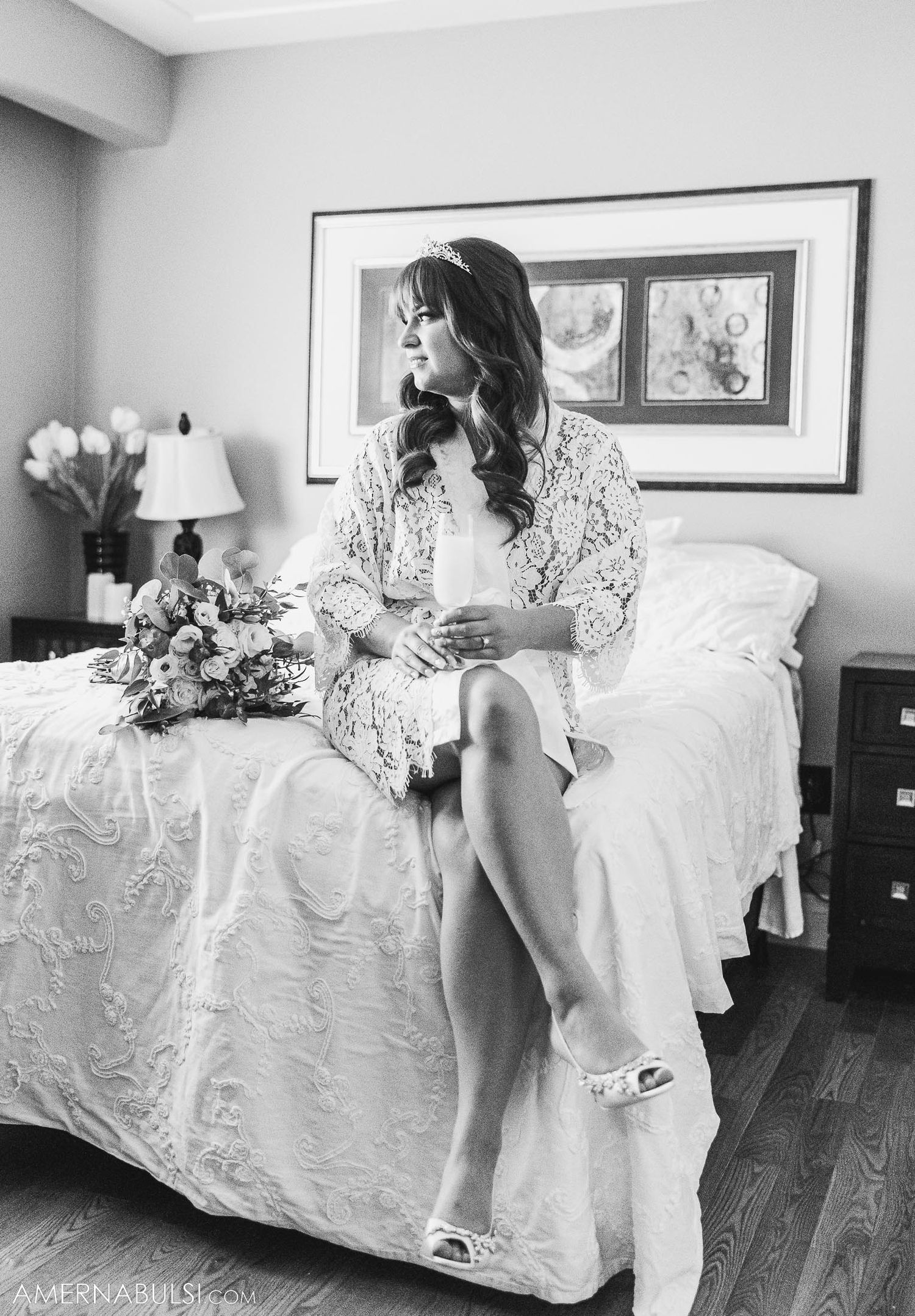 Black and white portrait of a bride in a lace robe sitting elegantly on a bed with flowers nearby.