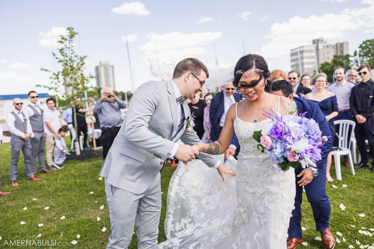 Newlyweds share a joyful moment on a grassy lawn with guests and city buildings in background.