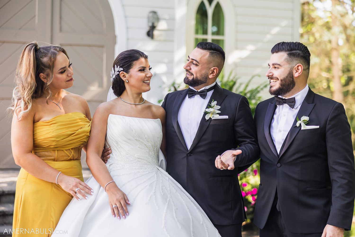 A wedding party of four stands together laughing outside a white chapel.
