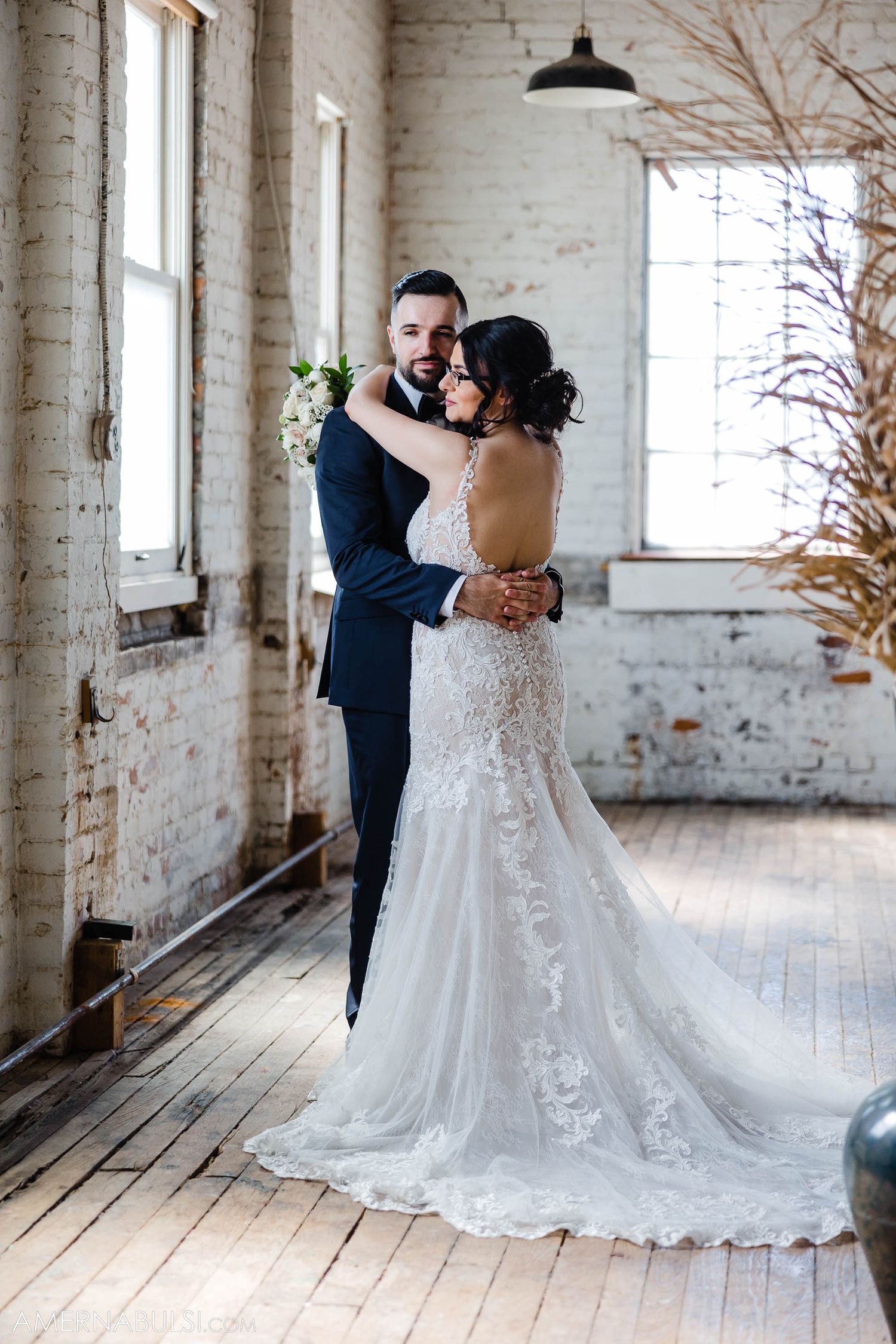 A romantic wedding portrait showing a couple embracing in front of bright windows in an industrial space.