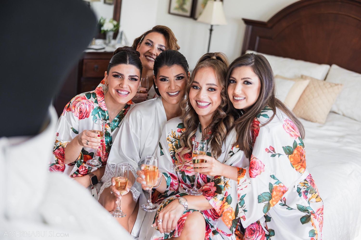 Wedding party in coordinated floral robes poses together on a bed while celebrating.