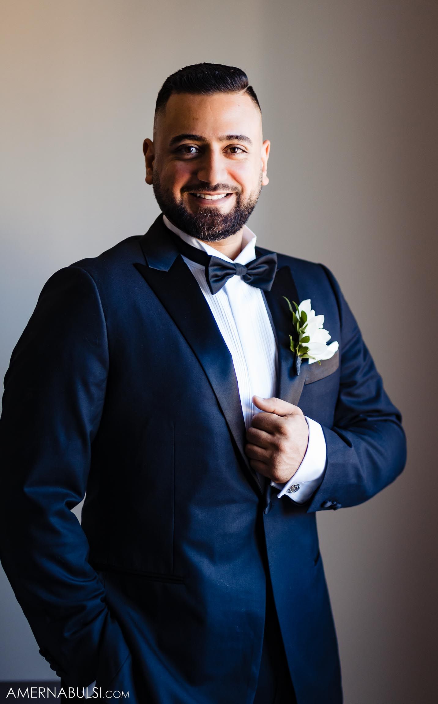 Groom in navy tuxedo and bowtie stands against grey background.