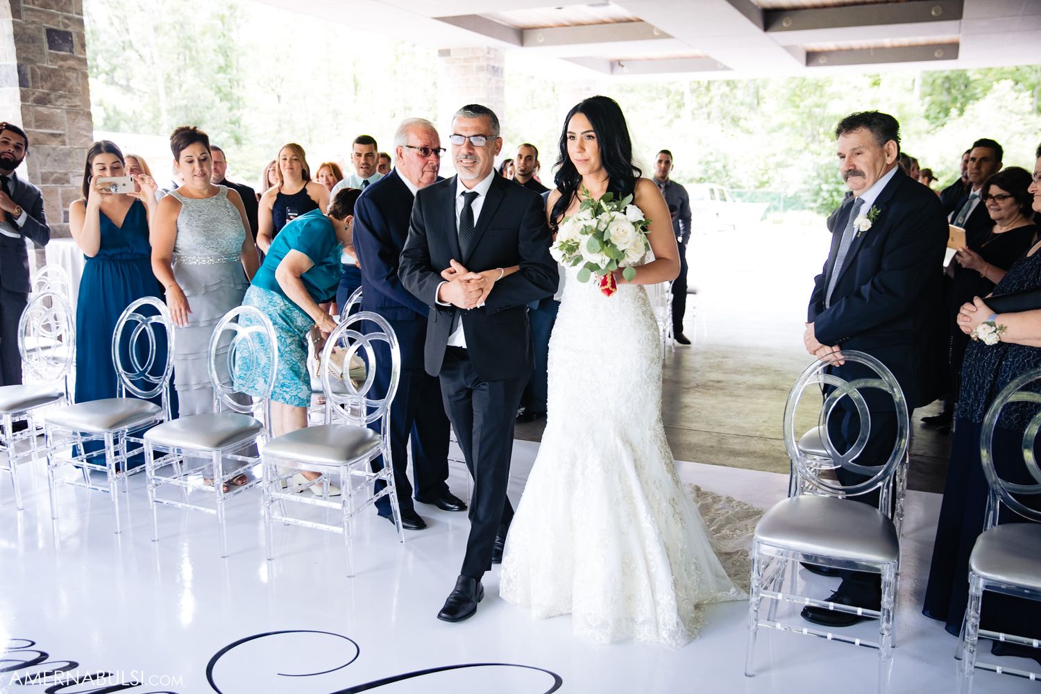 Wedding ceremony processional in modern venue with elegant ghost chairs and white floral arrangements.