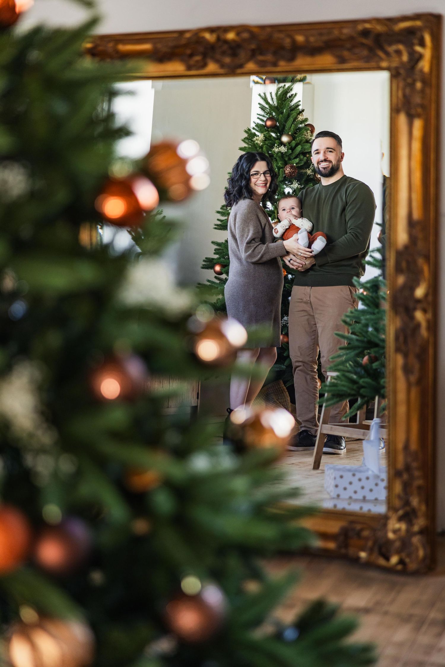 A holiday family portrait reflected in a gold-framed mirror with Christmas trees visible.