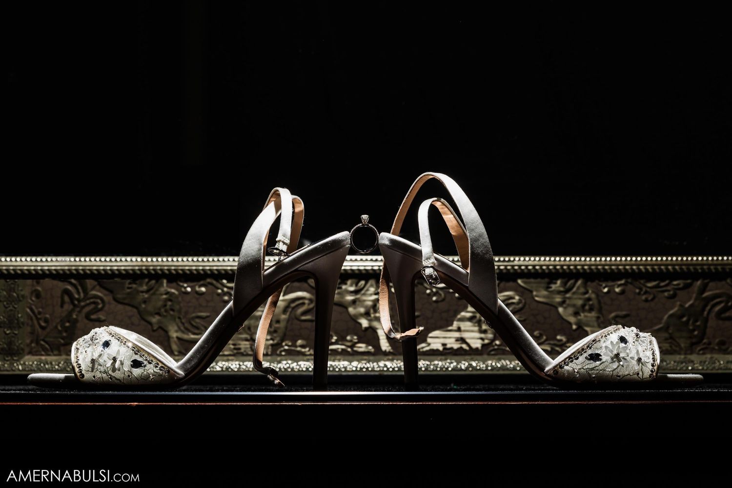 Dramatic lighting highlights elegant wedding shoes against ornate dark background.