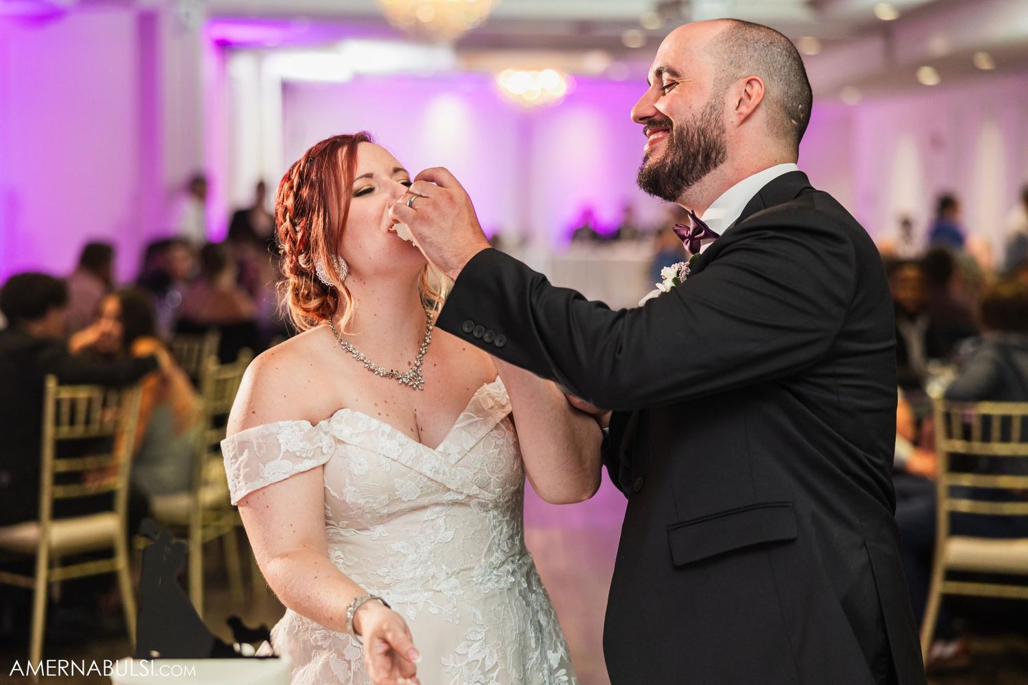 Couple playfully feeding each other wedding cake at reception with purple ambient lighting.