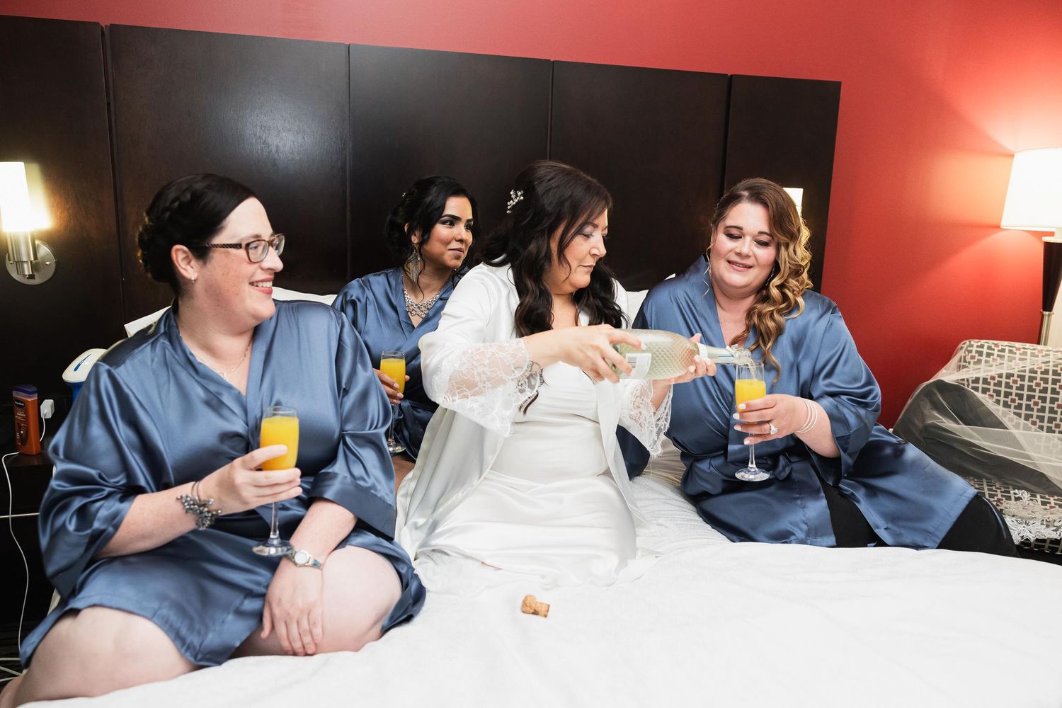 Wedding party in matching blue robes relaxes on bed while enjoying drinks.
